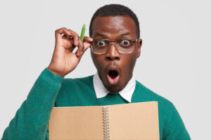 stupefied emotive dark skinned man opens mouth widely, holds pen, keeps hand on rim of glasses, hears shocking news, wears green sweater, isolated over white background. people and emotions.